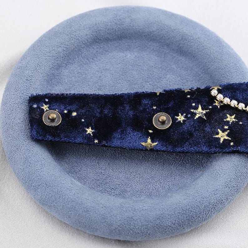 K&ouml;nnte beinhalten: Ein hellblaues Barett mit einem dunkelblauen Samtband. Das Band ist mit goldenen Sternen und kleinen Perlen verziert. Zwei bronzefarbene Kn&ouml;pfe befestigen das Band am Barett.