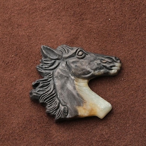 May include: A carved horse head pendant. The pendant is primarily dark gray with a cream and tan colored neck. The horse's mane is detailed with carved lines. The pendant is set against a brown background.