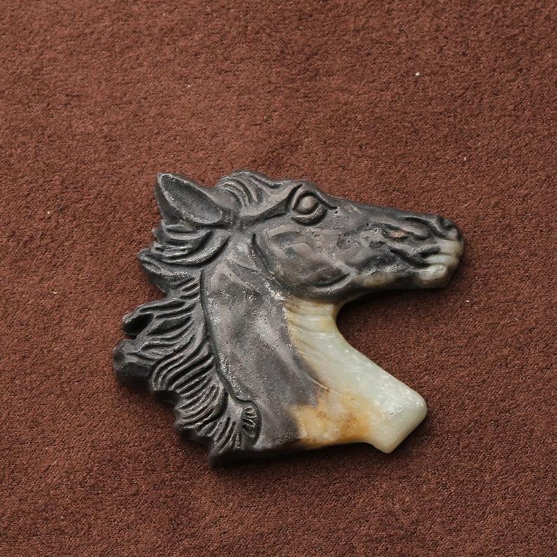 May include: A carved horse head pendant. The pendant is primarily dark gray with a cream and tan colored neck. The horse's mane is detailed with carved lines. The pendant is set against a brown background.