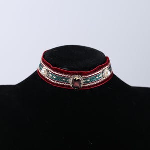 May include: A velvet choker necklace with a burgundy base and a woven green and white pattern. The necklace is adorned with a square, dark red gemstone in the center, and a pearl on each side.