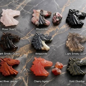 May include: A collection of carved horse head figurines in various gemstones, including rose quartz, obsidian, and cherry agate. The figurines are displayed on a marbled surface, with labels identifying each stone type. The horse heads are detailed and polished.