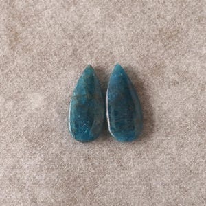 May include: Two teardrop-shaped, polished gemstones in shades of teal and blue. The stones have a marbled appearance with darker blue veining. The gemstones are displayed on a neutral, textured surface.