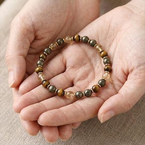 May include: A gemstone bracelet with a mix of brown, gold, and grey beads. The bracelet is displayed in open hands, highlighting the natural colours and textures of the stones. The bracelet is a piece of jewellery.