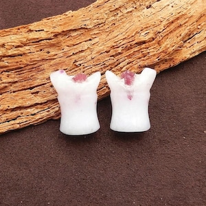 Puede incluir: Dos figuras de piedra blanca tallada, cada una con un acento de piedra preciosa rosa en la parte superior. Las figuras tienen forma de pequeños torsos. El fondo presenta un trozo de madera y una superficie marrón.