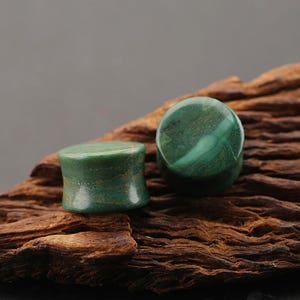 May include: Two circular ear plugs made of polished green stone with natural brown veining and a smooth, concave side profile. The plugs feature a mottled texture with varying shades of forest green and dark emerald, showcasing a natural stone finish.