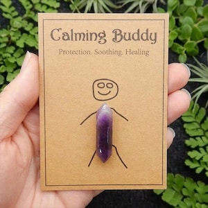 May include: A purple amethyst crystal on a brown card with the words "Calming Buddy" and "Protection. Soothing. Healing." The card has a simple drawing of a person. The crystal is held in a person's hand, surrounded by green plants.