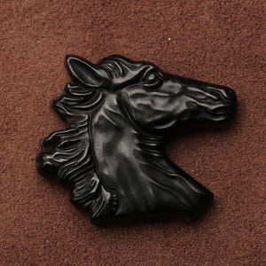 May include: A black horse head sculpture with detailed mane and facial features. The sculpture is made of a dark, possibly polished material, and is set against a brown background. The horse head is facing to the right.