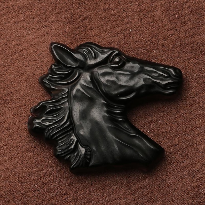 May include: A black horse head sculpture with detailed mane and facial features. The sculpture is made of a dark, possibly polished material, and is set against a brown background. The horse head is facing to the right.