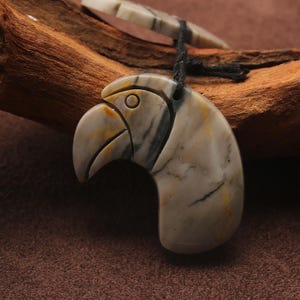May include: A carved stone pendant in the shape of a bird's head, featuring a beak and eye detail. The pendant is primarily cream-colored with gray and yellow veining. It hangs from a black cord. A second, smaller stone piece is visible above.