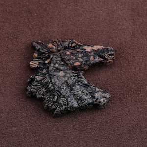 May include: A carved stone sculpture of a horse's head. The sculpture is primarily black with speckled orange and gray accents. The horse's mane and facial features are detailed. The sculpture is set against a brown background.