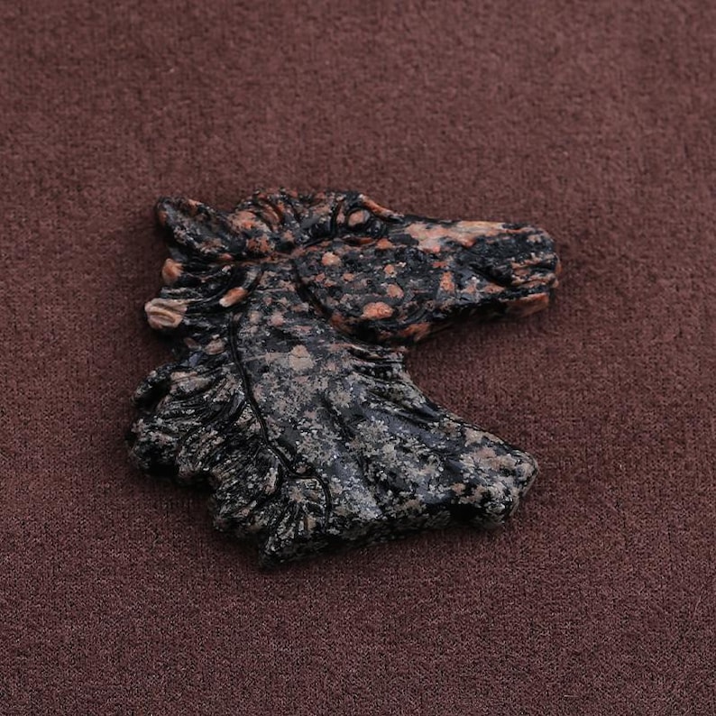May include: A carved stone sculpture of a horse's head. The sculpture is primarily black with speckled orange and gray accents. The horse's mane and facial features are detailed. The sculpture is set against a brown background.