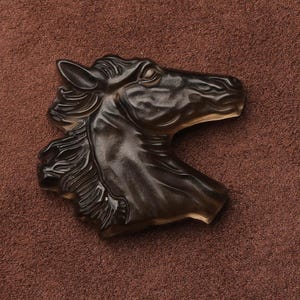 May include: A dark brown horse head sculpture with detailed mane and facial features. The sculpture is made of a translucent material, giving it a glossy appearance. The horse head is set against a brown background.