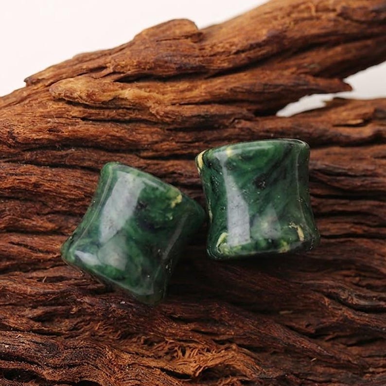May include: Two cylindrical ear plugs with a smooth, polished finish. The plugs feature a marbled pattern in deep forest green, emerald, and black tones with subtle lighter flecks throughout the material, creating a glossy, stone-like appearance.