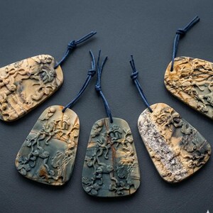 May include: Five carved stone pendants with blue cords. The pendants have a mix of beige, brown, and blue-green tones. Each pendant features a detailed relief carving of floral and organic designs.