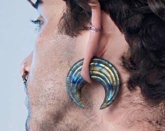 May include: A crescent-shaped ear weight crafted from polished labradorite stone, featuring iridescent blue and gold flashes. The jewelry is worn through a stretched earlobe, with a smooth, curved design that tapers to two pointed ends.