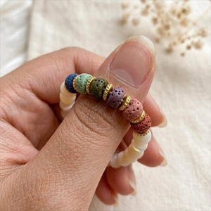 May include: A colorful beaded bracelet featuring a variety of textured, porous beads in shades of blue, green, brown, purple, and red, separated by gold-colored spacers. The bracelet is strung on a white, iridescent cord.