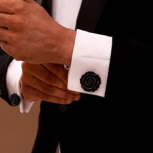 Peut inclure: Une paire de boutons de manchette en forme de rose noire fixés sur les poignets mousquetaires blancs d'une chemise. Les boutons présentent un design détaillé à pétales superposés dans une finition noire unie, contrastant avec le tissu blanc.