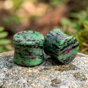 May include: A pair of cylindrical ear plugs crafted from polished stone, featuring a vibrant green base colour with distinct, irregular streaks of deep magenta and black mineral inclusions throughout the smooth, rounded surfaces of the jewellery.