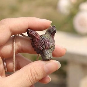 May include: A small, carved pendant shaped like a wolf head with textured fur details. The pendant features a blend of dark red, brown, and muted green tones and is attached to a thin, light-coloured, natural fibre cord for wearing as a necklace.