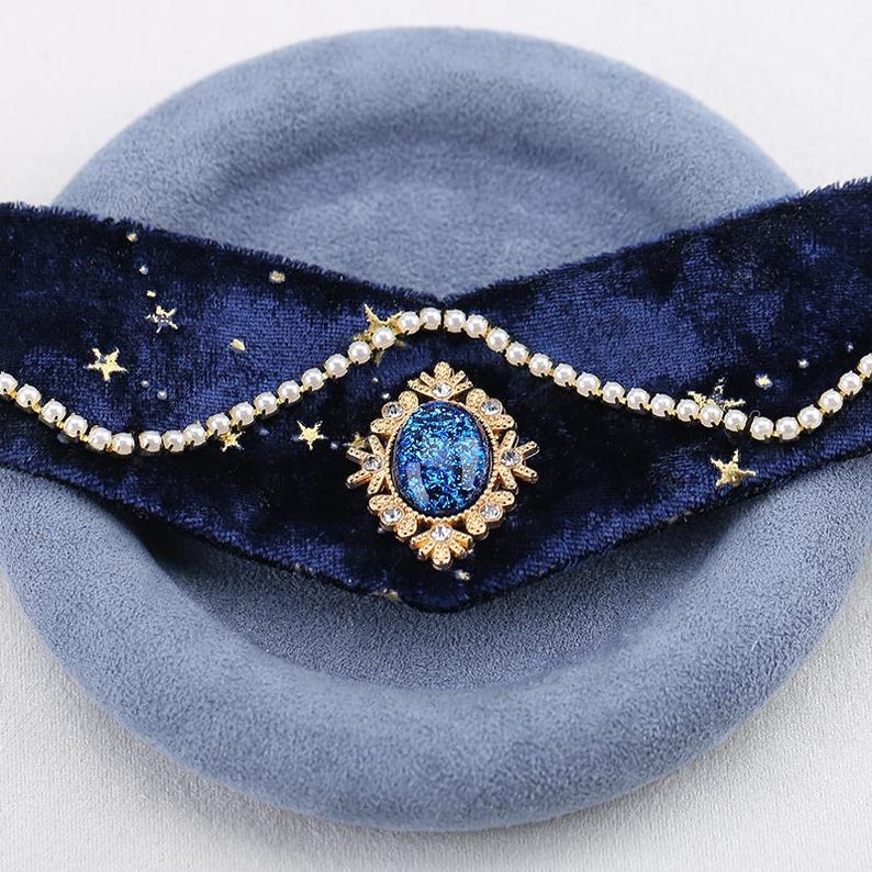 K&ouml;nnte beinhalten: Ein blaues Samt-Stirnband mit Himmelsdesign. Das Stirnband hat ein dunkelblaues Samtband, das mit goldenen Sternen und einem perlenartigen Besatz verziert ist. Ein zentrales goldfarbenes Juwel mit blauem Stein sorgt f&uuml;r Eleganz.