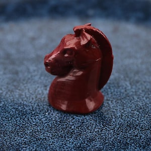 May include: A maroon-colored chess knight piece. The knight is carved in detail, showing the horse's head, mane, and neck. The piece is set against a textured blue background.