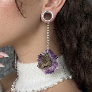May include: A silver ear gauge tunnel with a dangling chain attached to a raw, purple and brown amethyst crystal slice. The jewelry is worn in an earlobe, paired with a white pearl necklace featuring small silver beads.