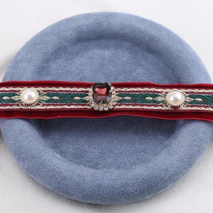 May include: A red velvet choker with a green and white embroidered design. The choker features a large, square, purple gemstone in the center, surrounded by small clear crystals. Two faux pearls are on either side of the gemstone.