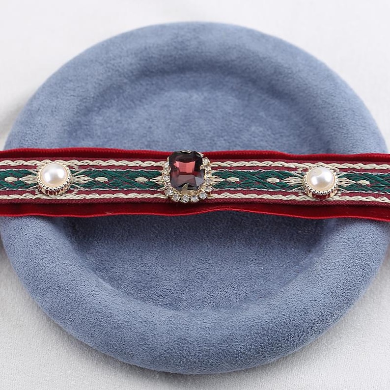 May include: A red velvet choker with a green and white embroidered design. The choker features a large, square, purple gemstone in the center, surrounded by small clear crystals. Two faux pearls are on either side of the gemstone.