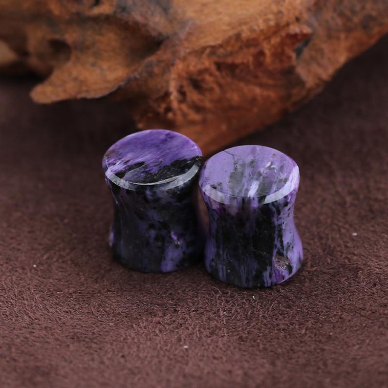 May include: A pair of cylindrical ear plugs crafted from polished stone, featuring a marbled pattern of deep purple and black streaks. The plugs have a smooth, glossy finish with a concave center, showcasing the natural variations in the stone's color.
