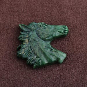 May include: A detailed, green carved horse head figurine. The sculpture is a profile view, showcasing the horse's mane, eye, and snout. The figurine is made of a solid, polished material, and is set against a brown background.