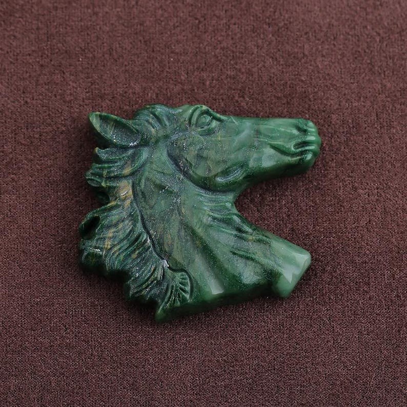 May include: A detailed, green carved horse head figurine. The sculpture is a profile view, showcasing the horse's mane, eye, and snout. The figurine is made of a solid, polished material, and is set against a brown background.