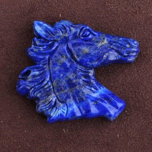 May include: A carved lapis lazuli horse head sculpture. The sculpture is a deep blue color with gold flecks, and the horse's features are detailed. The horse head is a bust, and the background is a solid brown.
