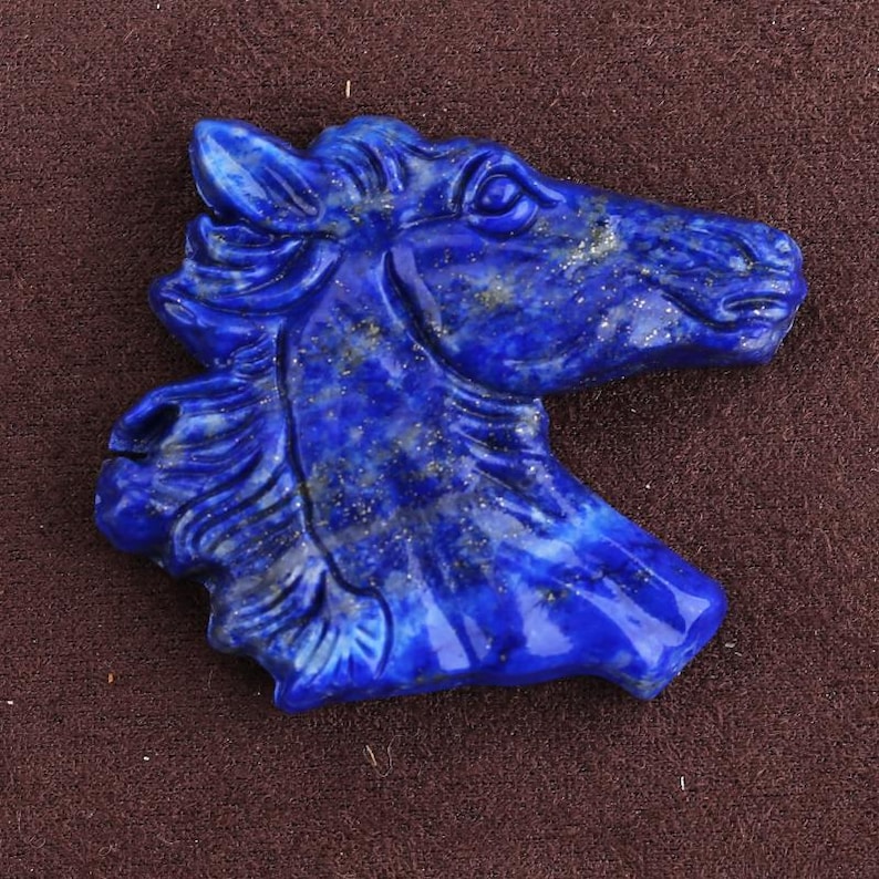 May include: A carved lapis lazuli horse head sculpture. The sculpture is a deep blue color with gold flecks, and the horse's features are detailed. The horse head is a bust, and the background is a solid brown.