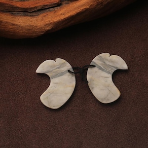 May include: Two gray and white marble Gua Sha tools connected by a black string. The tools have a curved, wing-like shape. The background is a dark brown color.