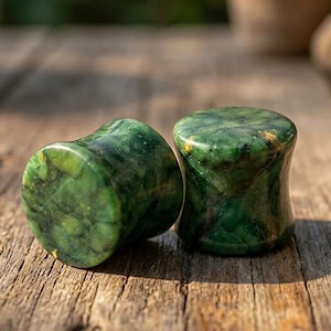 May include: A pair of cylindrical ear plugs crafted from polished green stone with natural dark veining and subtle gold-colored flecks. The plugs feature a concave hourglass shape with smooth, rounded edges, showcasing a deep emerald and forest green marbled pattern throughout the stone surface.