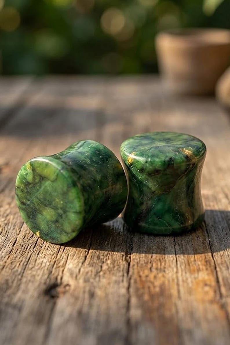 May include: A pair of cylindrical ear plugs crafted from polished green stone with natural dark veining and subtle gold-colored flecks. The plugs feature a concave hourglass shape with smooth, rounded edges, showcasing a deep emerald and forest green marbled pattern throughout the stone surface.