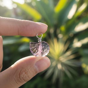 May include: A small, translucent, seashell-shaped pendant with a silver-coloured wire loop. The pendant is a light purple colour and is held between two fingers against a blurred green background.