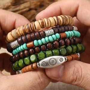 May include: A stack of wooden bead bracelets in various colors. The bracelets include light brown, dark brown, turquoise, and green beads. Some bracelets have silver-colored metal accents. The bracelets are held in a person's hands.