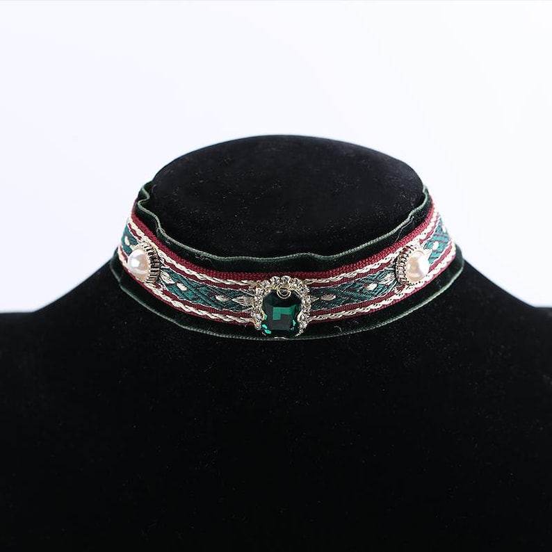 Pode incluir: Um colar gargantilha decorativo com um padr&atilde;o tecido verde e bord&ocirc;. O colar apresenta uma grande joia verde esmeralda no centro e dois pequenos detalhes perolados em cada lado. O colar &eacute; exibido em um manequim preto.