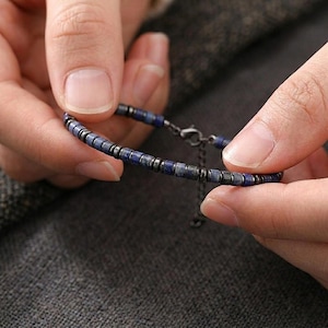May include: A beaded bracelet featuring small, dark blue and gray beads. The bracelet is held between two hands, showcasing the design and clasp. The beads are evenly spaced, creating a simple yet elegant accessory.