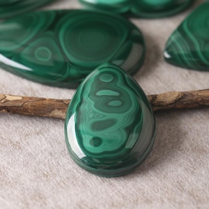 May include: Close-up of polished malachite gemstones in various shapes. The gemstones display a rich, deep green color with swirling patterns. One teardrop-shaped stone is in focus, with others blurred in the background. A small twig rests near the stones.