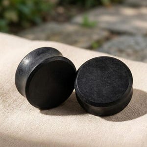 May include: A pair of solid black circular ear plugs with a smooth, matte finish. These cylindrical body jewelry pieces feature a flat front surface and a slightly recessed center, showcasing a consistent dark tone and a simple, uniform design.