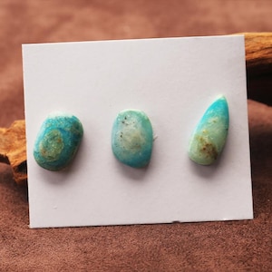 May include: Three turquoise gemstones of different shapes are presented on a white card. The stones display a mix of blue and green shades with distinctive patterns. These gemstones are likely for jewellery making or display.