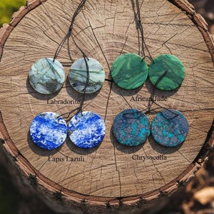 May include: Four pairs of round gemstone pendants on black cords. The gemstones include Labradorite, African Jade, Lapis Lazuli, and Chrysocolla. The pendants are displayed on a textured, brown wooden surface. Each pair is labeled with its gemstone type.