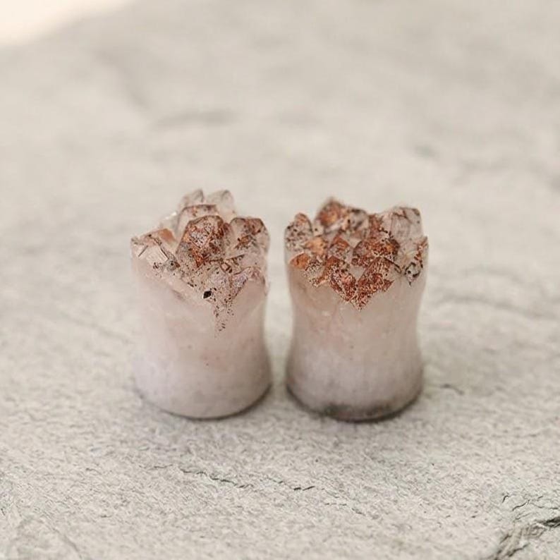 May include: A pair of ear plugs featuring a cylindrical shape with a flared base and a top surface composed of raw, crystalline structures. The material is a pale, translucent off-white stone with prominent reddish-brown mineral inclusions.
