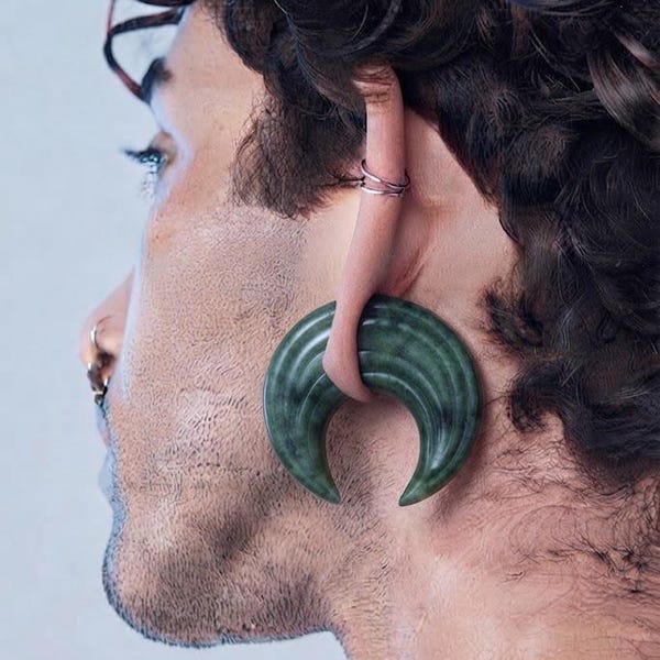 May include: A dark green crescent-shaped ear weight made of polished stone with subtle horizontal grooves. The jewelry is inserted through a stretched earlobe, featuring a smooth, curved design that tapers to two pointed ends at the bottom.