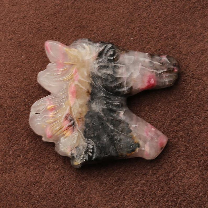 May include: A carved horse head figurine. The sculpture features a detailed mane with pink and yellow accents, a black and gray body, and a pink muzzle. The figurine is set against a brown background.
