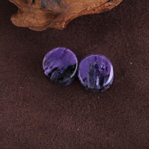 May include: A pair of circular ear plugs crafted from polished charoite stone, featuring a vibrant purple base color with distinct, swirling black and white mineral inclusions throughout the smooth, glossy surface of each individual piece.