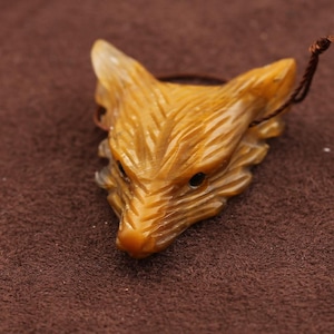 May include: A carved fox head pendant in amber. The pendant has dark eye details and a brown cord. The fox head is intricately carved to show fur texture. The amber colour is warm and inviting.