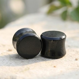 May include: Two solid black ear plugs with a smooth, polished finish. Each piece features a cylindrical shape with slightly flared edges on both ends, designed for ear stretching. The surface of the plugs is reflective and uniform in color.